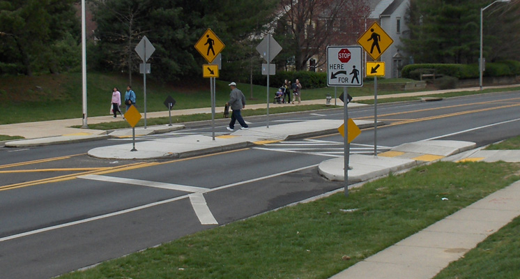 photo shows a mid-block crosswalk of a 5-lane street.  The sidewalk on each side of the street extends one lane into the street (bulbouts).  There is an island in the middle of the street, covering the width of one lane.  The crosswalk from each side is only one lane wide from the bulbout to the refuge island, and it is well marked with 3 signs facing each direction, each with arrows pointing to the crosswalk and indicating pedestrians (one has a picture of a stop sign and says 'stop here for pedestrians').